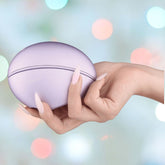 A manicured hand holds the Svakom Pulse Galaxie Air Pulse Stimulator in Metallic Lilac, highlighting its enticing suction effect against a softly blurred background with various colored lights.